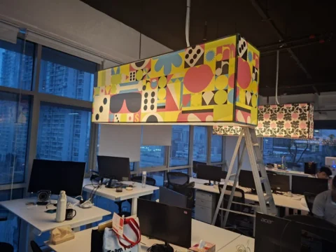 Finished office lighting by Best Volt, coloured geometric pendants lit over the desks with the city beyond a glass facade.