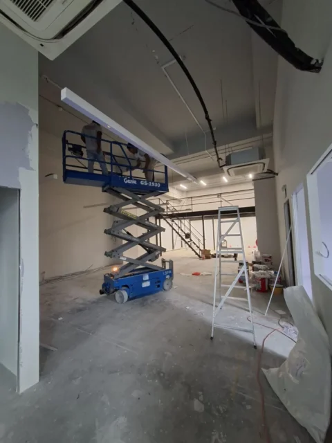 Genie GS-1930 scissor lift raised to a linear light, mezzanine stair behind, during a Best Volt high-access lighting installation.