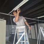 Linear lighting track being fixed from an A-frame ladder on a black ceiling, scissor lift behind, by Best Volt.