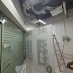 Gypsum partition with joint-compound patches and service openings, ladder and glass shutter in frame, a Best Volt first-fix shot from an F&B unit.