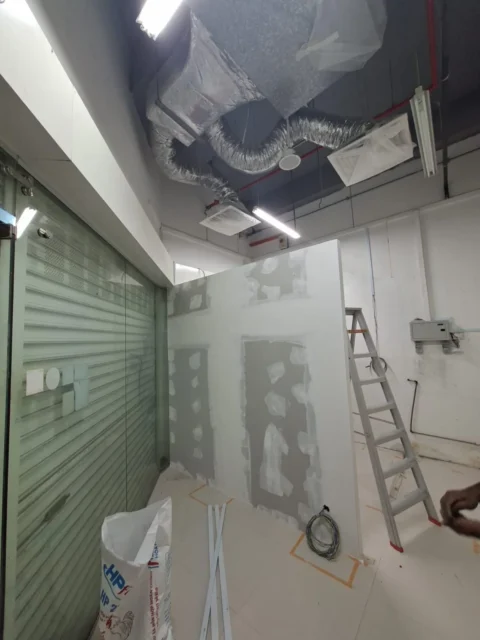 Gypsum partition with joint-compound patches and service openings, ladder and glass shutter in frame, a Best Volt first-fix shot from an F&B unit.